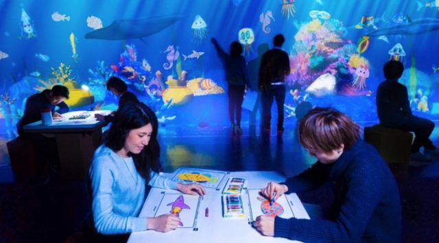 画像: お絵かき水族館 / Sketch Aquarium チームラボ, 2013 この水族館は、みんなが描いた魚たちが泳ぐ水族館です。 「紙」に自由に魚の絵を描きます。すると、みんなが描いた魚と共に、目の前の巨大な水族館で、その魚たちが泳ぎ出します。自分達の描いた魚に触ることもでき、触られると魚は、いっせいに逃げ出します。エサ袋に触ることによって、魚にエサをあげることもできます。 www.team-lab.net