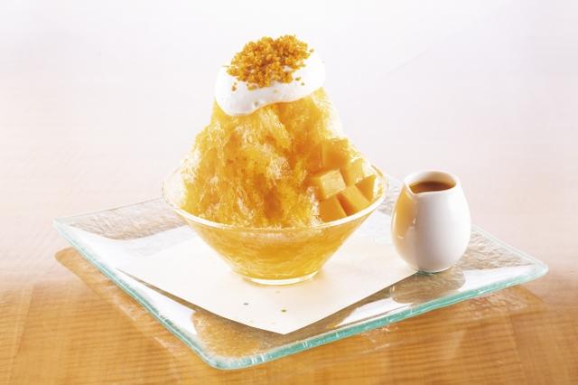 画像: 旬のフルーツ、マンゴーの美味しさ五感で楽しむ“マンゴーづくし”のかき氷です。氷の中にマンゴーアイス閉じ込め、食べやすくカットしたフレッシュマンゴーの果肉との調和を感じていただけます。別添えのソースは、マンゴーピューレとシロップを合わせ、甘さと酸味の黄金バランスを最大限まで引き出すために、じっくりと煮つめ薫り高いソースに仕立てました。仕上げに生クリームと細かくカットしたドライマンゴーをトッピング。 食感はもとより、オレンジ色のグラデーションが目にもあざやかで夏らしい色合いの氷スイーツはトロピカルなひとときを呼び込んでくれます。ホテルシェフ考案ならではの手間隙とアイデアを凝縮した氷デザートをぜひお楽しみください。