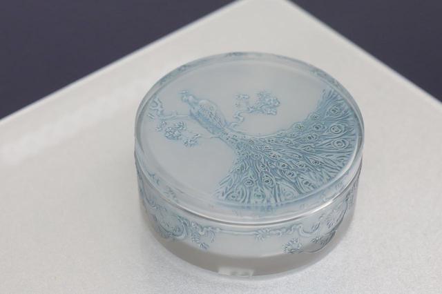 画像: ルネ・ラリック 円形蓋物「孔雀」1910年 金型のプレス成形で蓋は裏側から孔雀の姿を浮き彫りにしてサティネ加工で曇りガラス状にし、裏側からパティネで着色。器部分は浮き彫りが外側で内側をサティネ、外側からパティネで彩色している。ラリックにとってその後トレードマーク的な技術・技法となるものをすべて注ぎ込んだ作品で、そのガラス工芸作家としての公式デビューとなった「第6回装飾美術家サロン」で発表され、ラリック社の商品カタログに1番として登録された。 北澤美術館蔵