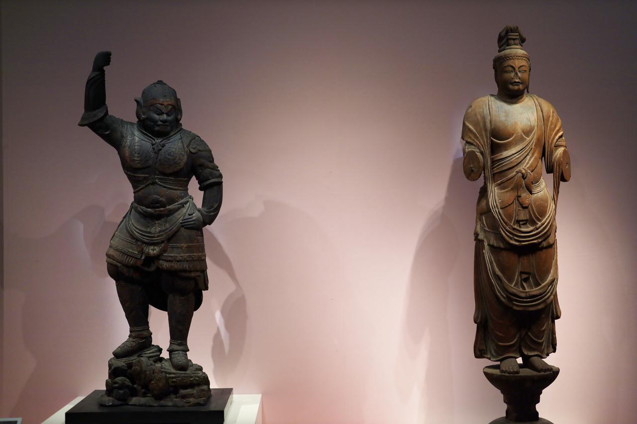 画像: 展示風景、左は鑑真に伴われて来日した唐の仏師の作と思われる唐招提寺の伝獅子吼菩薩立像（国宝）と、右は平城京で屈指の大寺だった大安寺に伝来する9体の奈良時代の木彫仏の一体、伝多聞天立像（重要文化財）で、鑑真のもたらした唐の技術を学んだ日本の仏師の作と思われる。
