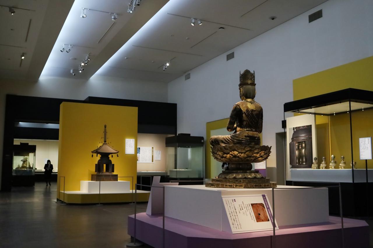 画像: 展示風景、叡尊坐像、叡尊が崇敬した舎利容器二件、叡尊が造らせた五智如来を表す鉄宝塔と、五智如来の中央・大日如来（運慶作）が一直線に並ぶ。運慶の大日如来の後ろには、台座に墨書された銘文と現代語訳のパネル。