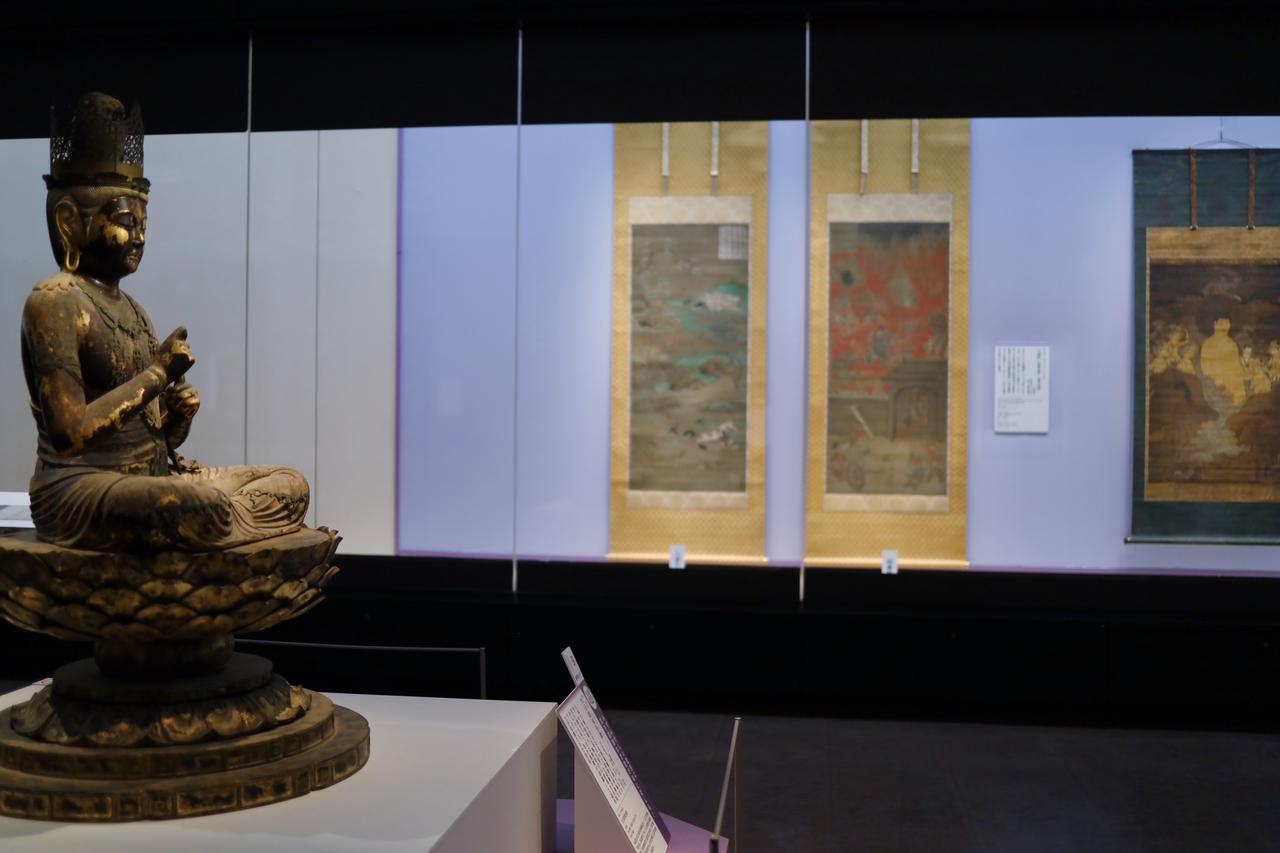 画像: 展示風景、運慶の国宝・大日如来坐像（円成寺）の横に、現世での行いに応じて輪廻転生で生まれ変わる先々を描いた六道絵（鎌倉時代・13世紀、聖衆来迎寺、国宝）と、阿弥陀如来の来迎が大自然の風景と一体化した山越阿弥陀図（鎌倉時代・13世紀、京都国立博物館、国宝）が並ぶことで、展示室内に曼荼羅の世界観が構成されている。大日如来坐像の後ろには、六道の思想を表現した絵巻物の地獄草子（奈良国立博物館）と餓鬼草子（東京国立博物館）が並ぶ。