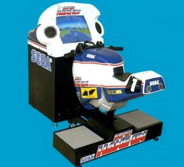 画像: こちらは1987年に発売の続編、「SUPER HANG-ON」(スーパーハングオン)です。初代は車体にディスプレイが埋め込まれていたので操作が重かったのですが、こちらは改良されて操作性が向上していました。なお、使われることのないフートレストは無くなってました(笑)。グラフィックは往年のロスマンズ・ホンダ風ですね。 static.giantbomb.com