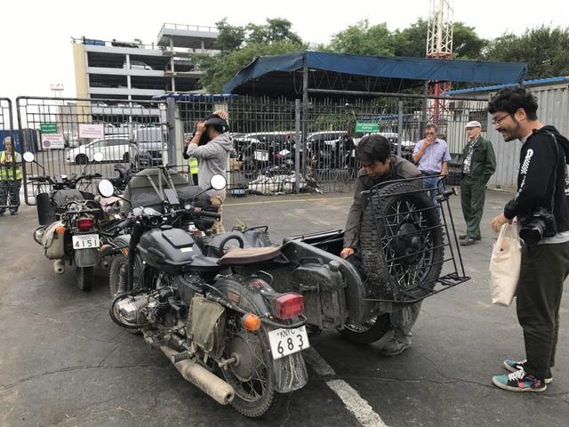 画像: それぞれのウラルを預ける前に、ヘルメットなどのライディングギアを側車のシートやトランクなどに収納。奥村カメラマン(右)はちゃっかり、自分の重い荷物をこのあと自分の手で持ち運ぶことを厭って、堀田さんに荷物を預けています・・・ワガママな人には困ったモノですね(苦笑)。