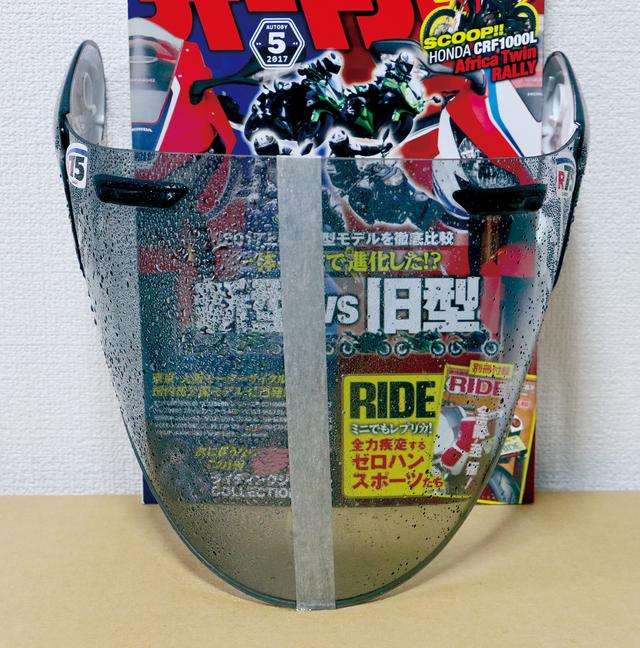 画像: シールド中央に貼ったテープの右側が施工面。写真は霧吹きで水をたっぷり吹き付けてから1分経過した状態。後ろに置いたオートバイ誌の文字からシールドコートZEROの強力な撥水性が判る。