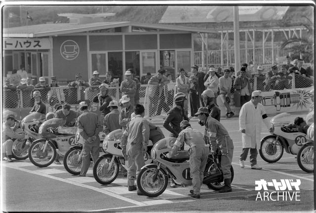 画像: 1964年日本GP・250ccクラス、スタート直前のグリッド。ポールを取ったジム・レッドマンのホンダRC165の向こうで、予選2位・ゼッケン22のヤマハRD56に乗るフィル・リードとレッドマンが言葉を交わしている。ゼッケン28のMZの右手にマイク・ヘイルウッドの姿があり、一番向こうで腕を組むのは2台目のRC165を駆る125ccチャンピオンのルイジ・タベリ。2列目手前、ゼッケン11のRD56のライダーは伊藤史朗。台数は少ないが豪華なメンバーだ。