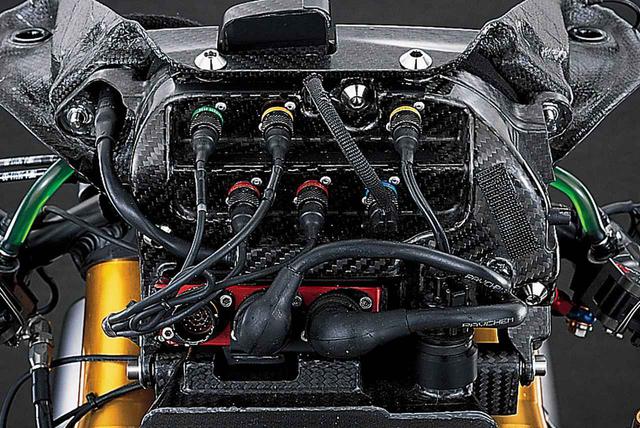 画像: メーターパネルを裏側(車体の正面)から見たところ。周辺の多くのパーツの取り付け基板となっているメーターパネルには、何本ものボルトを用いて強固にフロントカウル・アッパーステーが取り付けられている。マルチファンクションメーターの裏側に各種の信号線のコネクターが並んでいるが、これらはメーターに直接つながるのではなく、この部分がフロントまわりの信号線中継箱になっているものと思われる。向かって右のダクト上面にエアフローセンサーらしきものがマウントされ、そこからの信号線が上に伸びている。その左奥に見える赤い箱は、レーシングマシーンのデータシステムをサービスするドイツ・2D社製のデータロガー本体である。