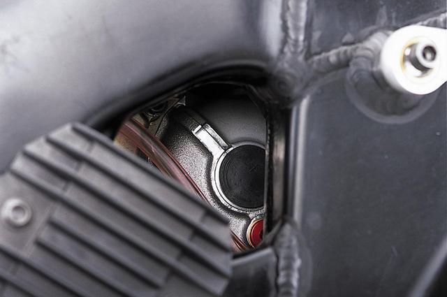 画像: エンジン右側前方の眺めは複雑だ。ラジエターの下にあるオイルクーラーへの配管は、カートリッジ式オイルフィルターの基部から出ており、オイルポンプからクランクケース内を通って圧送されたオイルは、まずオイルクーラーに入り、そこから戻った後にフィルターを通過してエンジン各部に向かっているものと思われる。注目すべきは、2本のエグゾーストパイプに取り付けられたO2センサーからの信号線カプラーの上部にある黒いプラグ(上下2本のボルトで固定)だ。何らかの軸加工をした跡なのは明らかで、ちょうどクランクケースの上下分割面にあり、ここを中心とする円形の張り出しが見えることから、こちら側にもバランサーが存在するのではないかと想像できる。