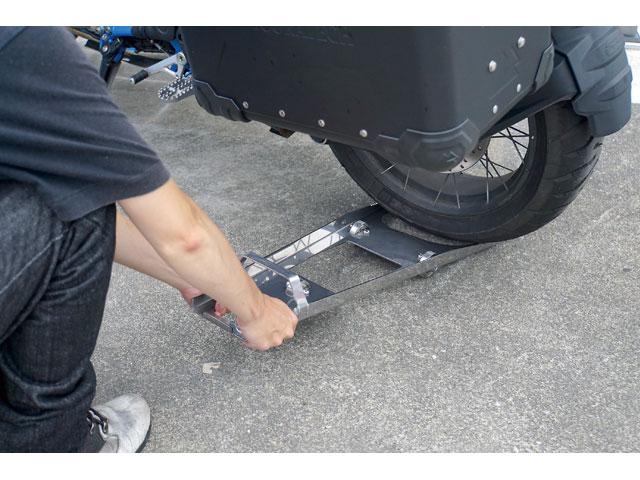 画像: 次に「バイクムーバープログレス」をリヤタイヤの下に差し込む。このとき先端のツノが確実にかかっていることを確認し、上から踏み込むとタイヤが浮く。そのままバイクの車体を押すと前輪を支点に弧を描くように動く。