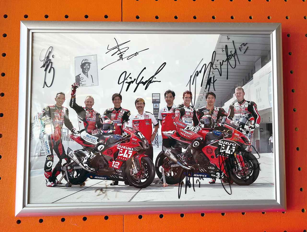 画像: ▲辻本さんが営むPILOTA MOTOの壁に飾られている一枚の写真。これはヨシムラ創立60周年となる2014年、ヨシムラは2008年以来となる2台体制で鈴鹿8時間耐久ロードレースに参戦。そのうちの1台は「ヨシムラ創立60周年メモリアルチーム レジェンド・オブ・ヨシムラスズキ」としてゼッケン12を付け、辻本聡／ケビン・シュワンツ／青木宣篤の布陣で出場した。