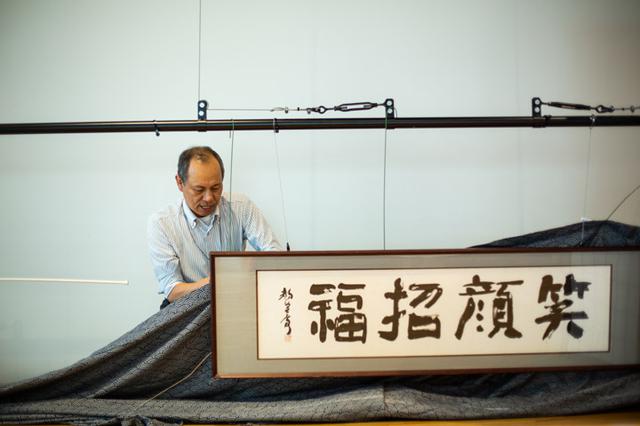 画像: 「笑顔招福(‶福"は旧字体)」の書は、日本ユニシス内のかつての部活動「書道部」の先生にお願いしたもの。幕を張り額の位置を整える松嶋安隆さん