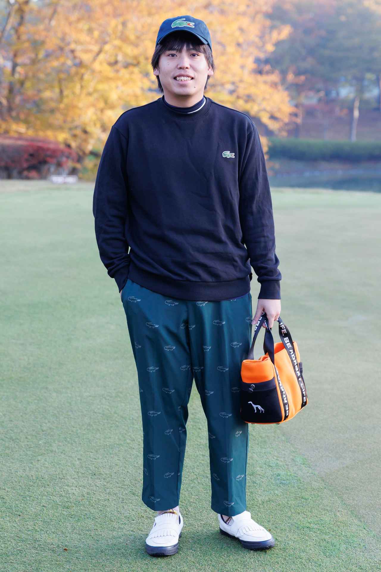 画像: ワニ柄が散りばめられたグリーンの総柄パンツを黒のスウェットシャツが引き立てているコーディネイト。オレンジのカートバッグや白のクラシカルなシューズも個性をプラス