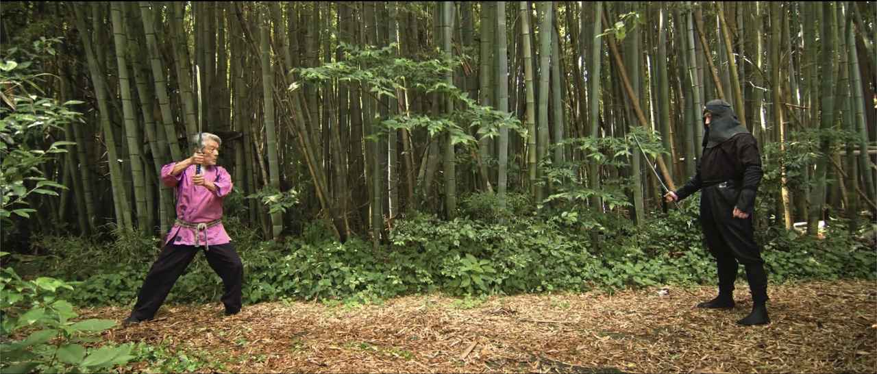 画像: 「緑の竹林にはピンクの道着が映えるんじゃないか？」と思いつき、自分で染め粉を買ってきて道着を染めたという倉田。この柔軟な発想、精神こそが若さの秘訣なのか。コロナ禍で先行きが見えなかった時代に、アクション魂に突き動かされたかのように弟子たちを相手に大暴れする倉田先生は必見！　忍者姿は中村浩二監督。