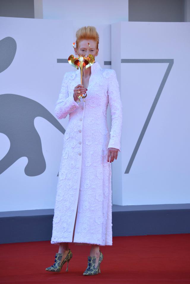 画像: ヴェネツィア国際映画祭にて、仮面舞踏会風のゴールドのマスクを持ってフォトコールに応じるティルダ。