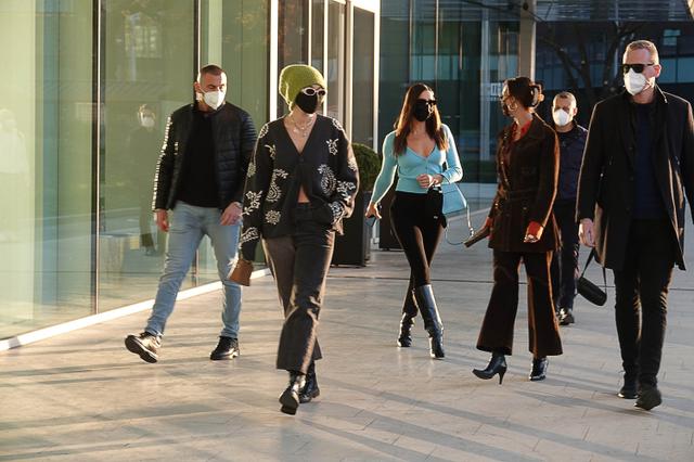 画像: ミラノ市内を移動中のジジたち。1人につき1人ずつボディガードらしき屈強な男性がついているのが印象的。柄カーディガンにグリーンのニット帽がジジ、ブルーのトップスがイリーナ、パンツスーツがベラ。