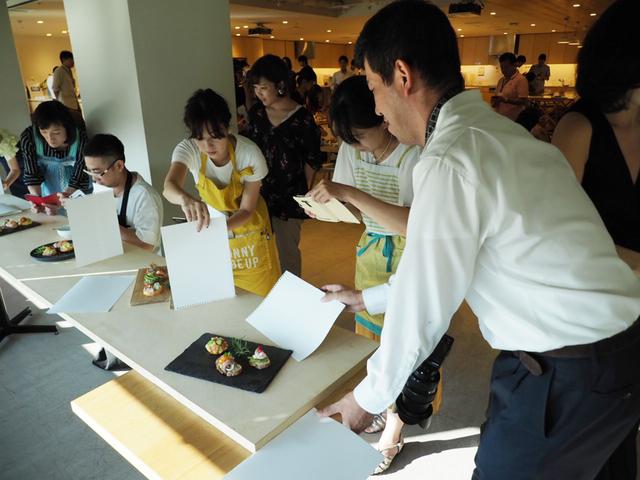 画像: 白い紙を即席のレフ板代わりに自然光が入る窓際でおのおのが作った自慢の料理を撮影。「ここはレブ板屋の僕に任せてください!」と思わずレフ板の角度を指導をするケンコー・トキナーの田原氏(写真手前)。