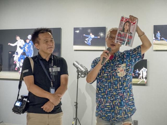 画像: 写真展初日の会場で挨拶をするカメラマン編集部・坂本。坂本が手にしているのはカメラマン10月号で、TOP LIGHTでも小城さんの作品が紹介されています。後ろにみえるのが今回使用したフラッシュ。