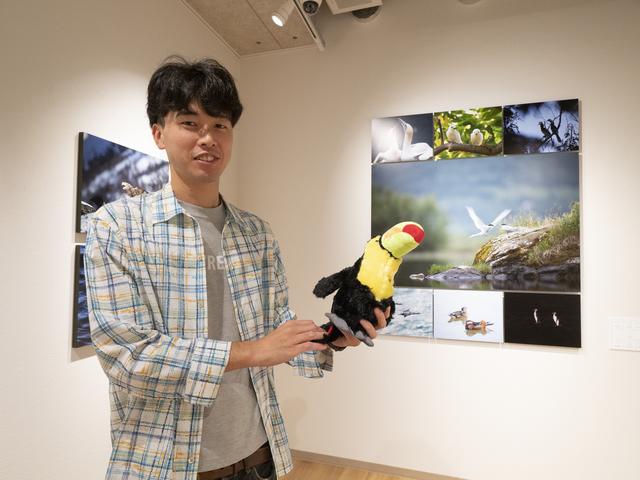 画像: 菅原貴徳さん。菅原さんが手にしているのはサンショクキムネオオハシのぬいぐるみ。残念ながら今回の展示にはこの鳥の写真はないという。