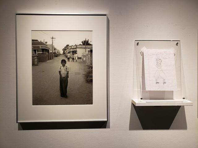画像: A.Rishaちゃんという少女の写真の横には、彼女が書いた絵とタミル語で書かれたノートの1片が並ぶ。そこには「私はこの村が好き。先生も好き。この村に原子力発電所が立って、危険と隣り合わせになったことは、たくさんの人に影響を与えている。」と書かれている。