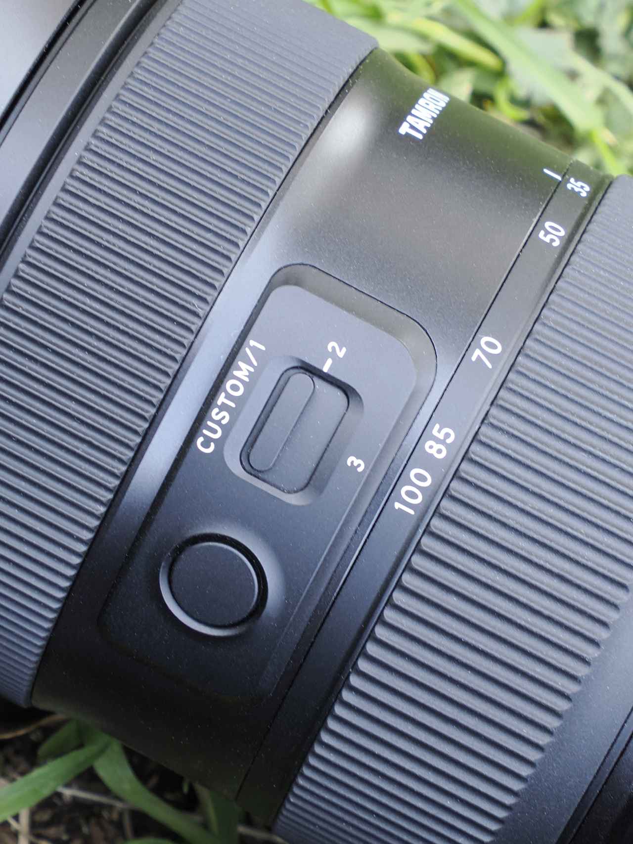 画像: 丸い「フォーカスセットボタン」は、カメラ側のカスタム設定で割り当てた機能を使うことができる。いっぽう、四角いほうのスライド式の「カスタムスイッチ」は、タムロンレンズ専用のソフトウェア「TAMRON Lens Utility」で設定する機能を割り当てられる。3つの機能を切り替えて使うことができる。割り当て可能な機能はソニーE用もニコンZ用もほぼ同じだが、少しだけ異なる機能もある。詳しくは「TAMRON Lens Utility」のメーカーサイトで。