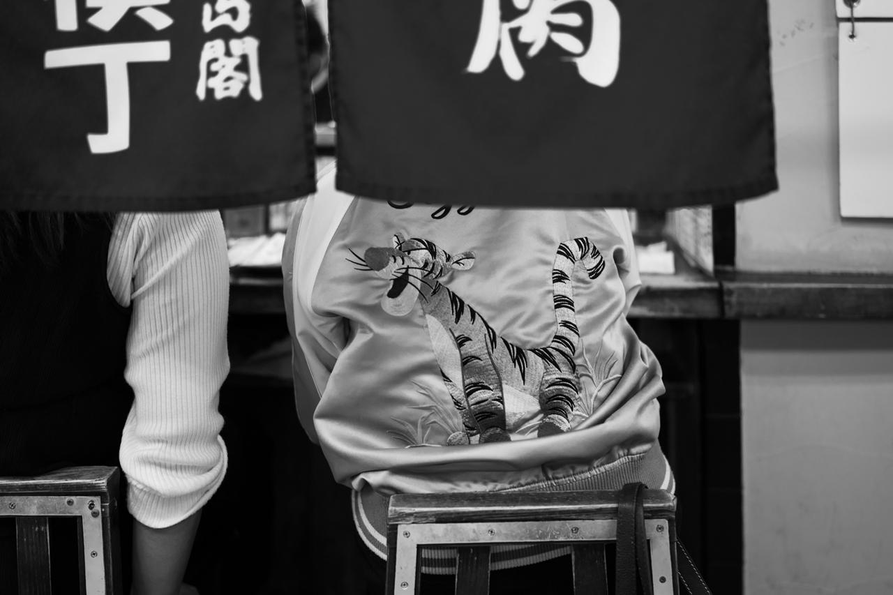 画像: 21時すぎの人工光での撮影で、ISO160という感度を維持できているのはF1.4という明るさのおかげ。スカジャン特有のサテン生地の光沢感が白トビせずに階調豊か、かつキャラクターの刺繍の立体感が際立っており、暖簾との素材感の対比がよく表現されている。 ■絞りF1.4　1/60秒　ISO160