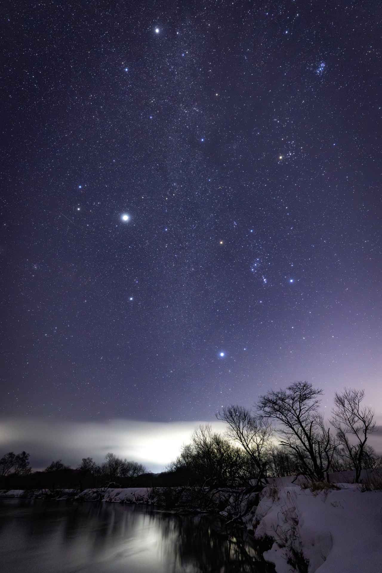 画像: 蛇行する釧路湿原の釧路川を前景に入れて、昇る冬のセブンスター（1等星）と木星を撮影した。画面上からぎょしゃ座のカペラ、牡牛座のアルデバラン、双子座のポルックス、オリオン座のベテルギュスとリゲル、こいぬ座のプロキオン、大犬座のシリウスだ。冬の賑やかな星座と風景をバランス良く網羅できるのがこのレンズの特徴。さらにF1.4という開放F値のおかげで、画面中央に淡く流れる冬の天の川がハッキリと浮かび上がる。F2.8では、再現不可能な暗い星まで捉えることができる。 ■絞りF1.4　8秒　ISO4000　WB:3600K　※釧路湿原