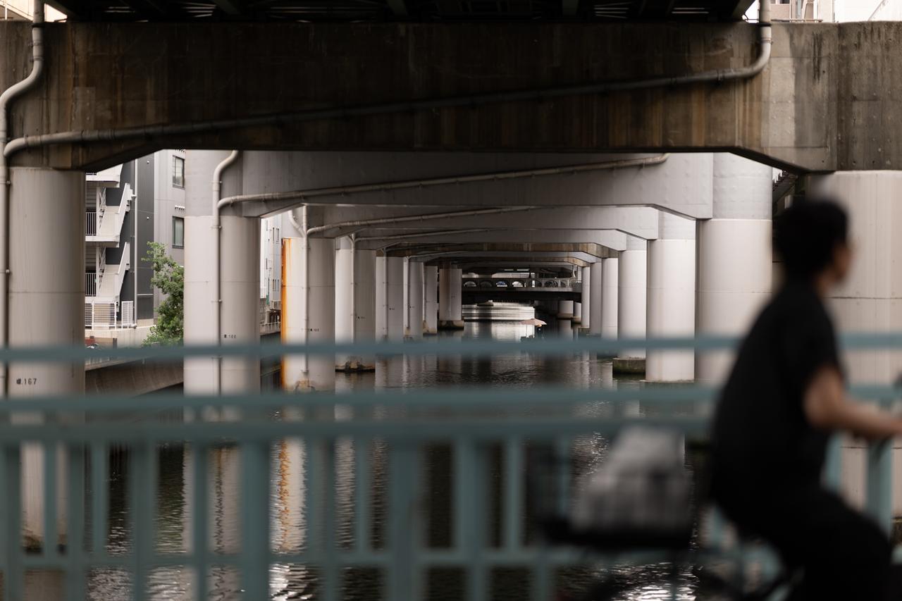 画像: 橋脚が連続する水辺の風景を、手前の自転車の人物をぼかしつつ撮影。奥行きのある構図でも画面の隅々までシャープに解像している。中央奥に小さく写り込んだ船頭が棹を漕ぐ姿まで、輪郭が際立っているのには驚かされた。 ■絞りF1.2 1/800秒 ISO100