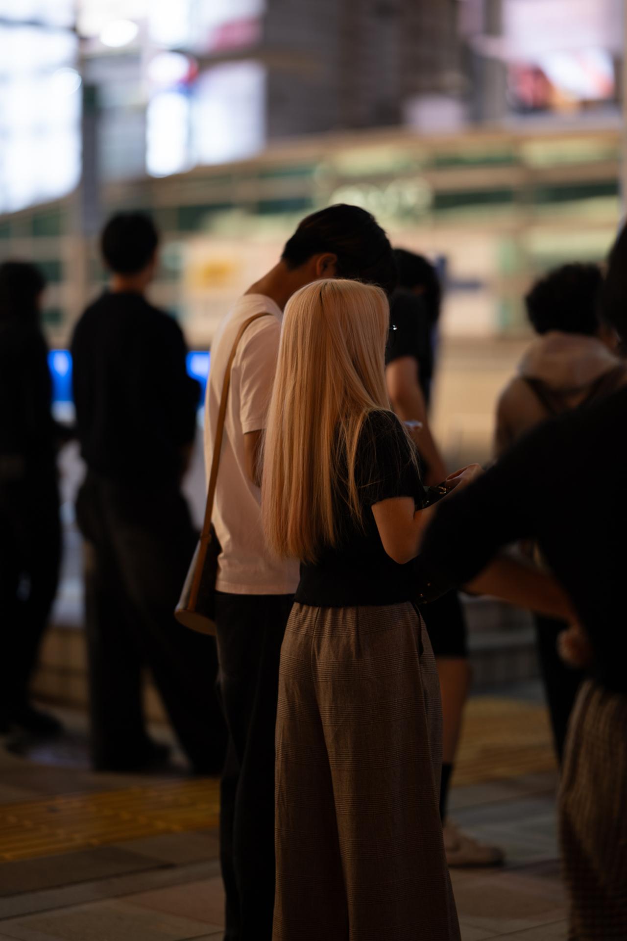 画像: 夜の街角、信号待ちをする人の群れの中から、長い金髪の後ろ姿を一枚。街あかりを受けた髪の艶が一本一本ほどけるように描写され、ハイライトも白トビせず、しっとりと粘っている。背景に並ぶ光は大きくやわらかなボケとなって溶け、人混みの中でも彼女だけが静かに浮かび上がる。ISO125のまま1/60秒で切れるのも、F1.2の明るさがあってこそだ。 ■絞りF1.2 1/60秒 ISO125