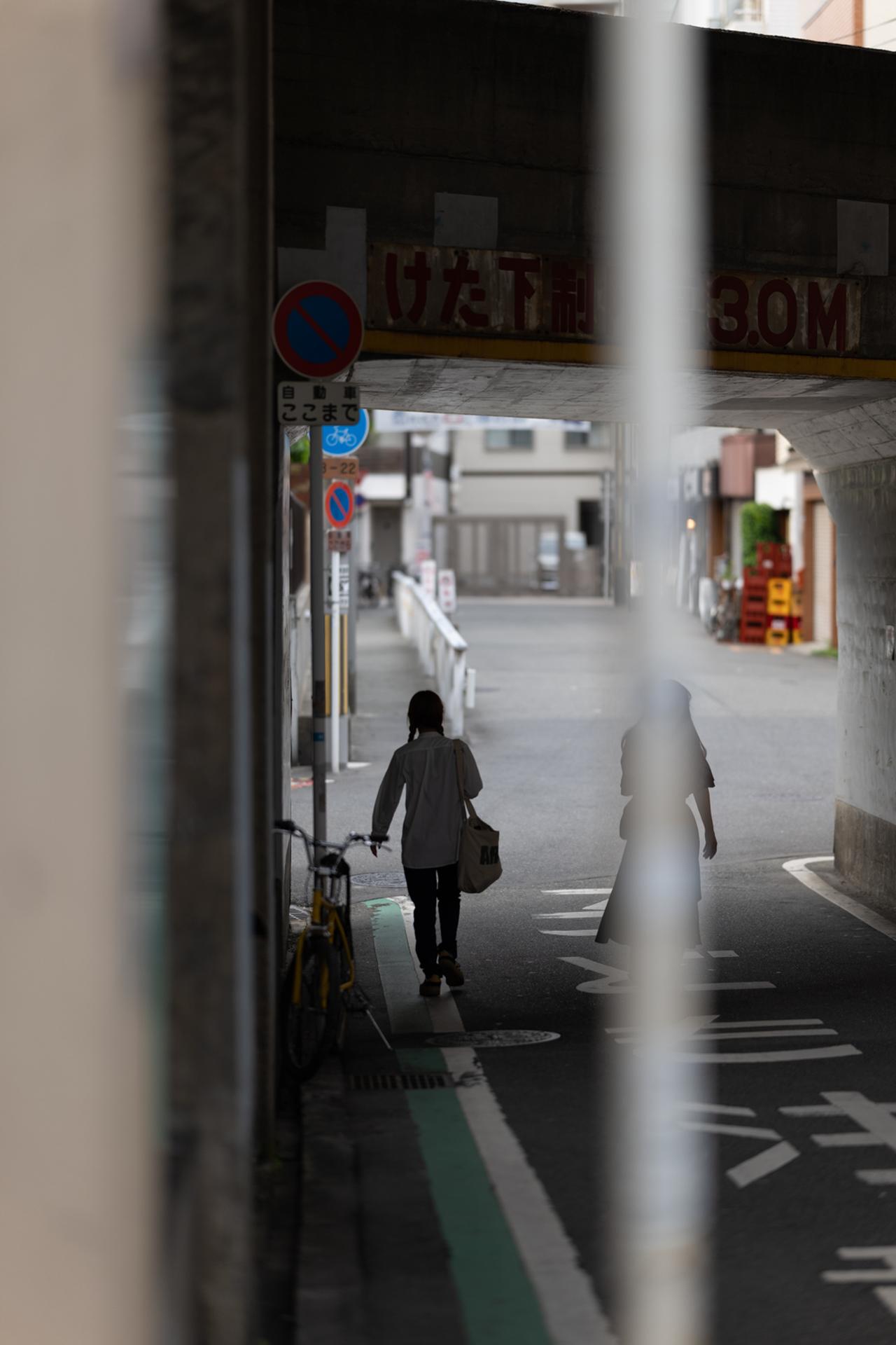 画像: 高架下、逆光気味の路地を歩く人影を開放F1.2で。手前のポールを表情の目隠しとして使いながら、奥の女性のシルエットが浮き出るようだ。軸上色収差の補正がしっかり効いているのか、逆光でも輪郭ににじみが出ない。層を重ねるように前景・被写体・背景を分離できるのは、この明るさならではの芸当ではないか。 ■絞りF1.2 1/1000秒 ISO100