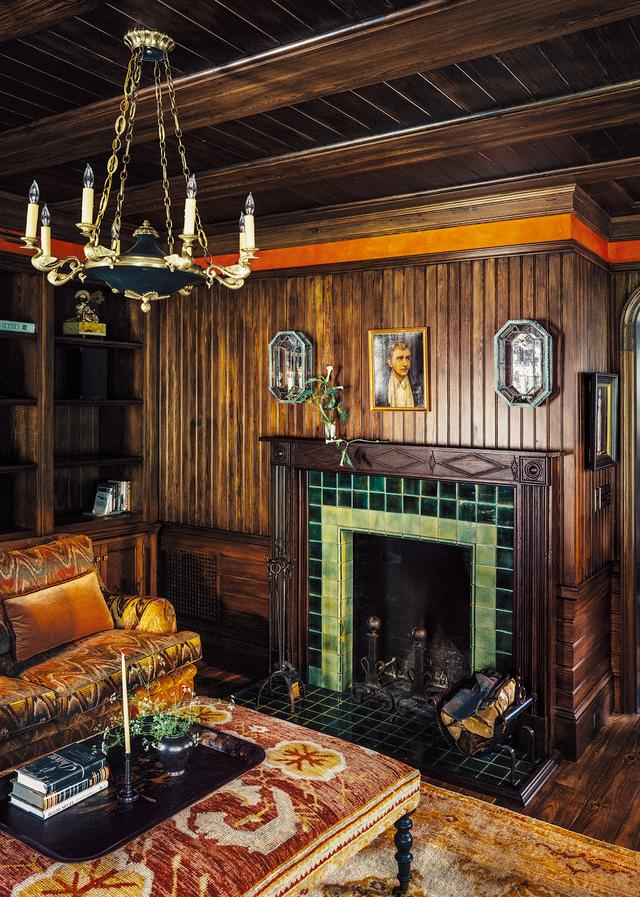 画像: シアタールームを灯すのはアンティークのエンパイア様式のシャンデリア。パイン古材で覆った壁にはポンペイレッドの漆喰のフリーズ(註:壁の帯状の装飾)と、1930年代の鏡つき燭台(NYのアンティーク家具店「Carlos de la Puente」で入手)があしらわれている。19世紀製の木製の暖炉枠は「バンタム・タイルワークス」特注の緑色のタイルをあしらい、その存在感をさらに際立たせている。「Watts 1874」の炎のモチーフを織り込んだベルベットでくるんだソファと、トルコのウシャク絨毯地で覆ったオットマンは、ともに「ローマン&ウィリアムス」によるデザイン。