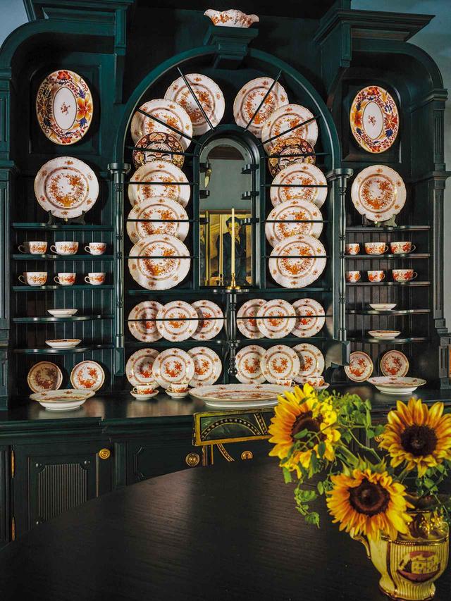画像: ダイニングエリアのディスプレイ用キャビネットは「ファイン・ペインツ・オブ・ヨーロッパ」の深いティール色が印象的。そこに並ぶのは「ヘレンド」が前世紀に発表した「インディアン・バスケット」柄のテーブルウェア・セット。アンティークのイギリス製ラスターウェア(註:光沢ある金属釉を施した陶磁器)のピッチャーとヒマワリがダイニングテーブルを鮮やかに彩る。