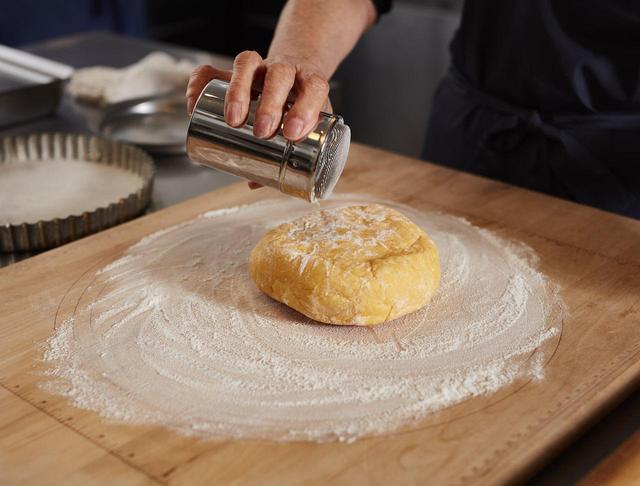 画像: １ タルト生地を作る。フードプロセッサーに薄力粉をいれ7〜8秒ほど回す。砂糖と塩を加えて3〜4秒ほど回す。冷蔵庫から取り出した冷たいバターを約1㎝の角切りにし、バターが小豆粒くらいの大きさになるようにパルスで回す。少し粉気がある程度で止める（この時回しすぎないようによく注意すること。フードプロセッサーにより回す時間は異なるのでよく注意して）。卵黄を1個づつ加えてその都度軽く回す（この時も回しすぎないように注意）。まとまったら取り出し、ラップで包んで冷蔵庫で１時間ほど休ませる。フードプロセッサーがなければ、同様の順番で手でこねても。台に打ち粉（分量外）をし、生地をおき、上にも小麦粉を振る。