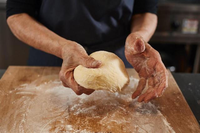 画像: 5 台の上に打ち粉をし、４の生地のおく。空気を抜き、手で平たく伸ばし、表面がツルツルになるように縁を丸め込む。丸めたら15分ほど休ませる。