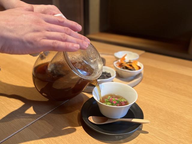 画像: さいの目切りした野菜が入った器に目の前で養生スープを注いで仕上げる季節のミネストローネ。スープに浮かんだ梅の花とシャクの葉も春らしい