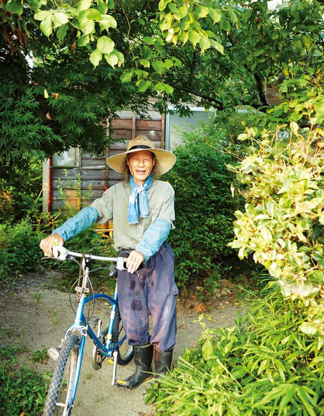 画像: 野良着スタイルの川口さん。木々や花が豊かな自宅の庭先で。車や機械類を持たない川口さんは、腰に鎌を差して、さっそうと自転車に乗って田んぼや畑へ向かう