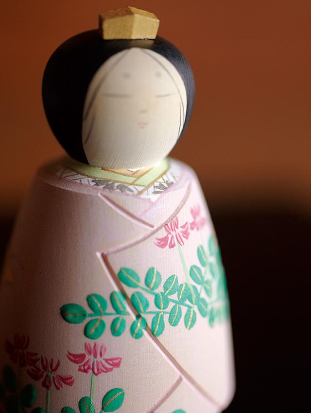 画像: 春の花、れんげを描いた雛人形。薄いピンクの着物地の奥にはやさしい木目が透けて見える。額に垂れた髪の毛がなんとも艶やか。柔らかな眉、すうっと一文字に引いた目、小さなおちょぼ口……と、柔和な顔立ちも「松尾雛」の魅力のひとつ