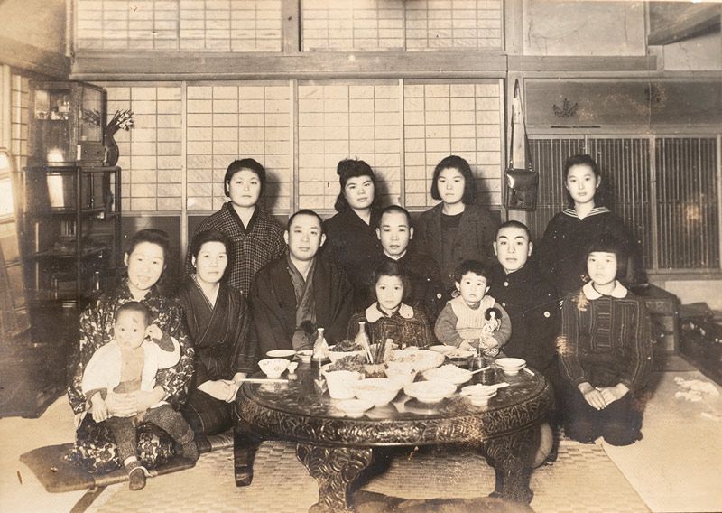 画像: 家での写真。前列左から2番目が母親、その右隣が父親。前列中央が多良さん、その右隣が8歳下の妹。制服姿の青年は「当時下宿していた海軍さん」