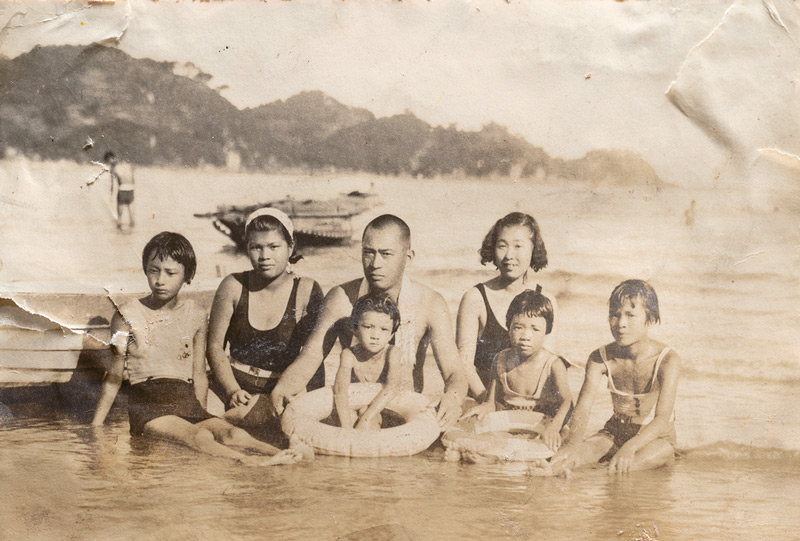 画像: 5歳くらいのころ、家族で海水浴に行ったときの1枚。後列中央の男性が父親、膝に座って浮き輪を手にしているのが多良さん。周りにいるのは姉たち