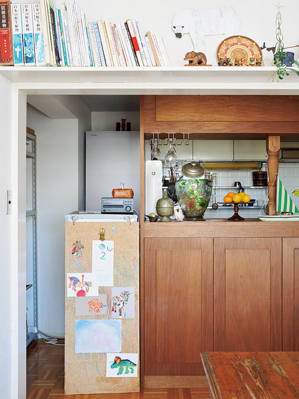 画像: 食卓から見た台所。イラストを貼ったコルクボードは、飲み物や粉類を入れているミニ冷蔵庫の側面隠しの役割も