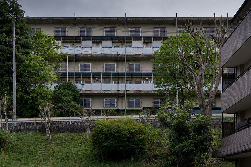 画像: こちらは団地の敷地の外からみたメゾネット住棟（多分N-32号棟）。敷地の起伏がわかりますでしょうか。こちらは2017年撮影。建て替えのためすでにメンテナンスがされておらず、かなり痛みが目立つ痛ましい姿になってしまっています