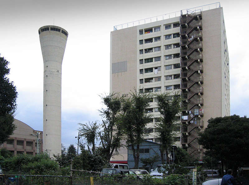 画像: 「桐ヶ丘団地」には1棟だけですが、14階建ての高層棟も建っていました。こちらは、昭和43年度建設。起伏に富んだ敷地のためか、給水塔も複数(おそらく3基)ありました。写真の左が給水塔です
