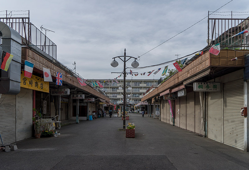 画像: 「桐ヶ丘団地」の商店街。後ろの住棟は4階建てではなく5階建て。実は昭和37年度に計画がブラッシュアップされ、以降は5階建ての建物も建てられるようになりました。すべてを確認できていませんが、5階建ての住棟には増築されてないものも見られるので、もしかしたら、それ以降の住棟にはお風呂がついてたのかもしれないですね