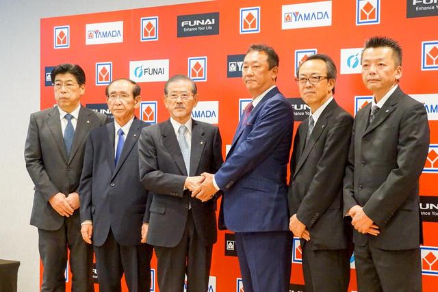 画像: 中央で握手しているのが、株式会社ヤマダ電機 代表取締役会長 兼 取締役会議長の山田昇氏(左)、船井電機株式会社 代表取締役 執行役員社長 船越秀明氏(右)。流通と製造の大手が、文字通りがっちりと握手している