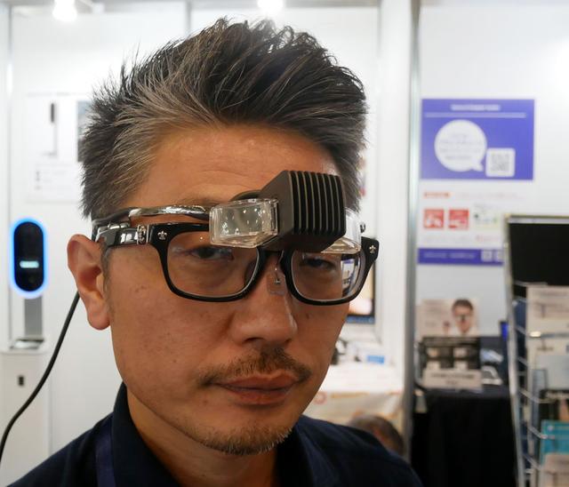 画像: ▲Enhanlaboブースでは、メガネスーパーのノウハウを投入したウエアラブルデバイス「b.g.」を展示(発売中)。オーバーグラスタイプ(眼鏡on眼鏡)で、両眼視、デバイスは有機EL、パネルは1280×720という仕様だ。グラス部分は移動させることができ、周囲を見ながら表示を見たり、使用しない時は上にはね上げておくことも可能だ。本体はモバイルバッテリーで駆動でき、HDMI入力に対応するので、スマホやタブレットからの動画再生も楽しめる