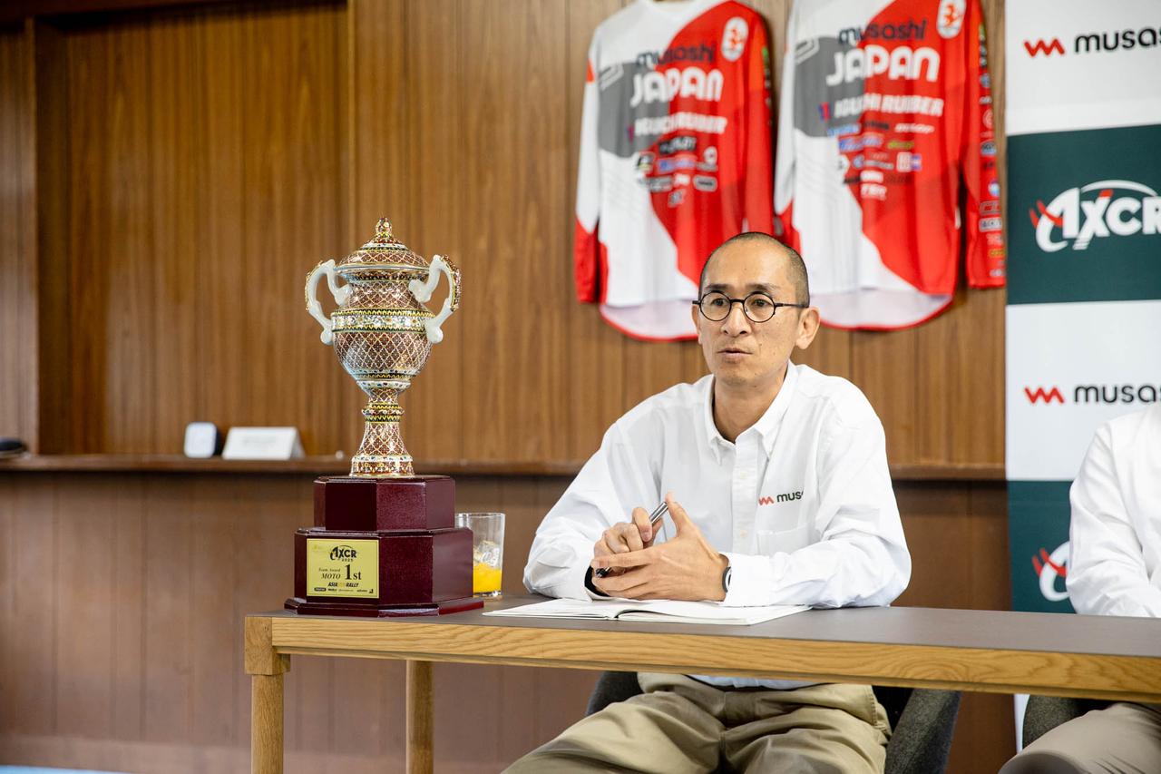 画像: 株式会社ムサシの岡本篤代表。後ろにかかっているジャージはISDEポルトガルの時のもの。2019年の日本代表トロフィーチームをバックアップし、釘村とのリレーションが生まれた。自身もオフロードバイクに乗る