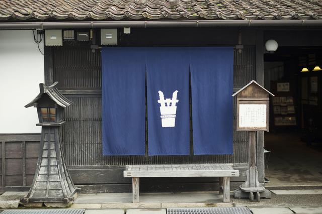 画像: 辻本店の「角樽」が描かれた暖簾。勝山では「暖簾のかかる町並みプロジェクト」で100を超える家々に業種を表すデザインの暖簾がかかっており、町歩きする私たちの目を楽しませてくれる