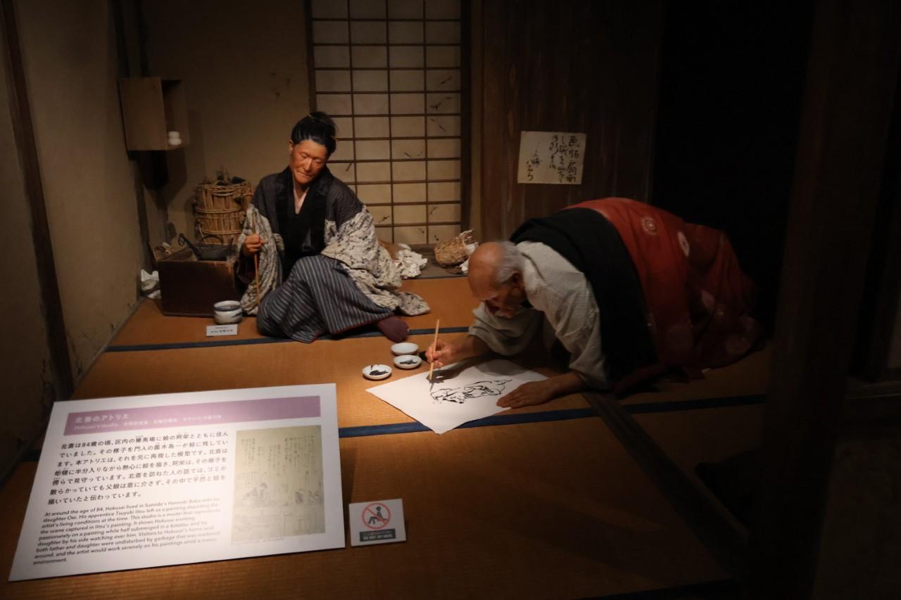 画像: 北斎のアトリエ再現模型。84歳の時の北斎の姿を再現した展示。娘の阿栄（おえい）と住んでおり、こたつに入りながら筆を持っていたという