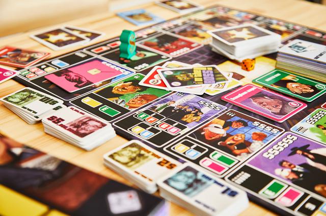 画像: ボードのマス目には、「A」「E」などの手持ちの人物カードを示すアルファベットが書かれている。アルファベットは商談を成立させる条件を示している。