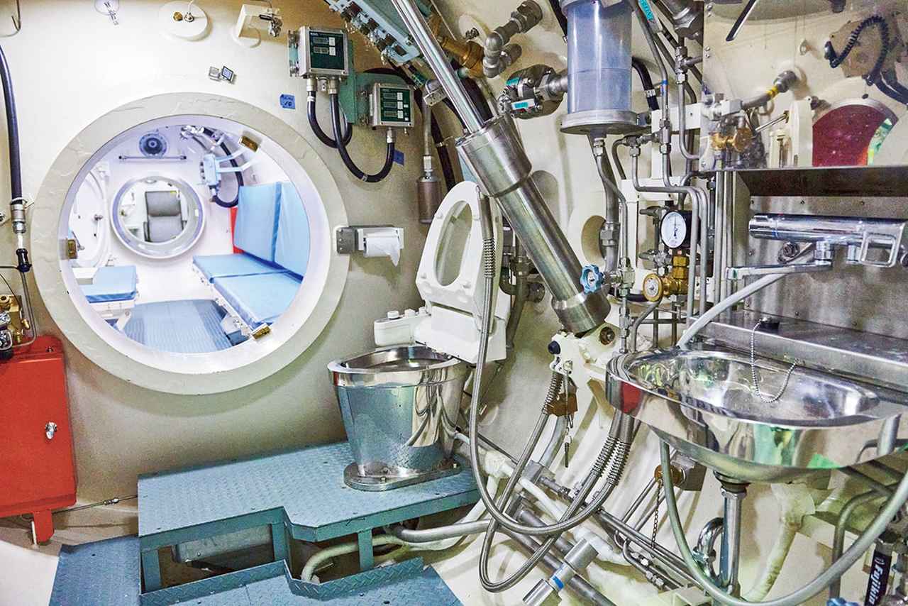 画像: 訓練で1週間過ごすという「飽和潜水用艦上減圧（再圧）タンク」。手前にシャワー、並んでトイレがあり、ここを使用するときはモニターも遮断される。奥は生活スペース