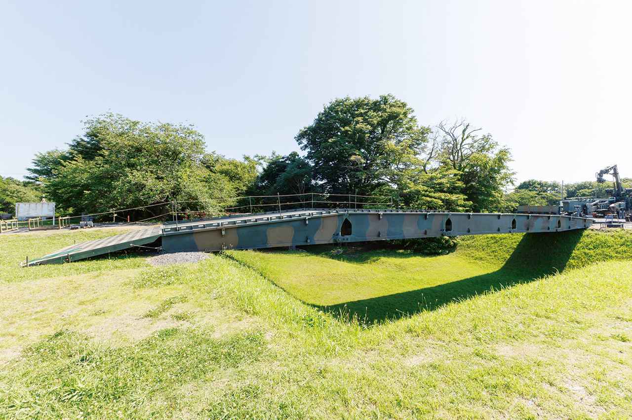 画像: 破壊された橋の代わりに、07式機動支援橋で架橋。架設車の対岸側にはスロープを設置し、川岸と段差ができないように高さを調節する